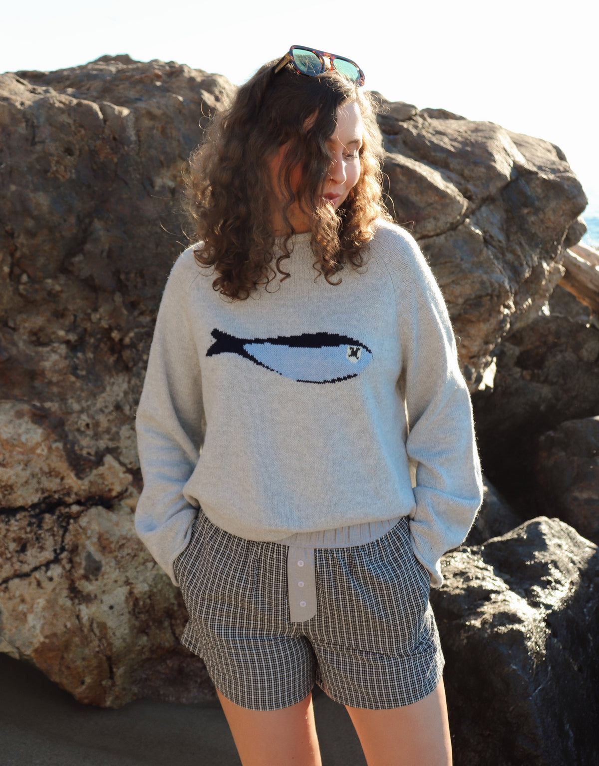 Woman outdoors by a rocky coastline, wearing a gray fish sweater and black shorts. 