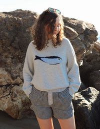 Woman outdoors by a rocky coastline, wearing a gray fish sweater and black shorts. 