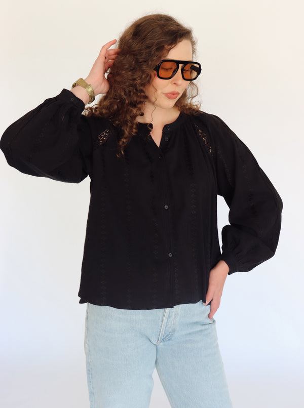 A woman wearing a black long-sleeve blouse with lace front and embroidered shoulder details, paired with light blue jeans and black sunglasses, posing against a plain white background.