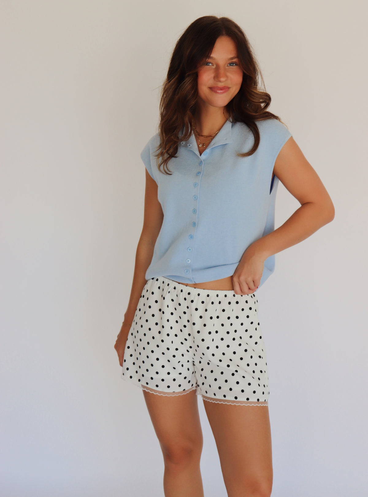 A woman modeling a light blue short-sleeve button-up shirt and white shorts with black polka dots, standing against a plain light background.