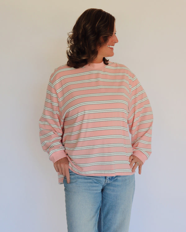 A woman wearing a pink long-sleeve crewneck shirt with thin white horizontal stripes, paired with light blue jeans, standing against a plain off-white wall.