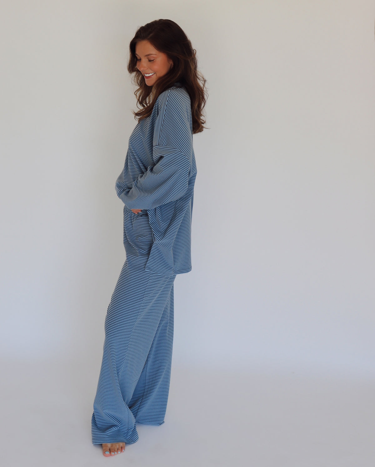 A woman modeling a matching blue striped lounge set, consisting of a long-sleeve top and wide-leg pants, against a plain white studio background.