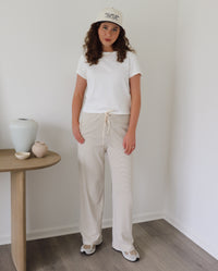 A person wearing loose, wide-leg drawstring pants in a cream, blue and beige stripe pattern, paired with a white top and white sneakers, standing in a minimalist room with wood floors.