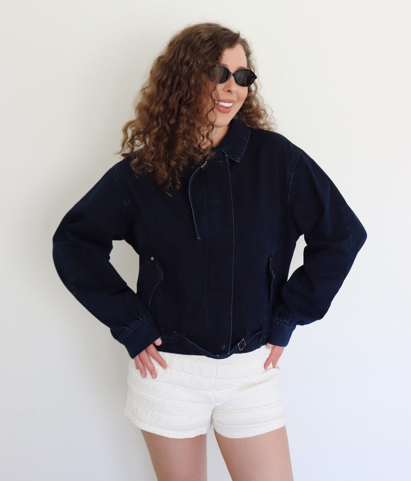 A woman with curly brown hair and sunglasses wearing a dark navy blue cropped denim jacket and white textured shorts, posing against a plain white wall.