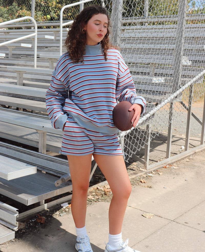 A woman stands on an outdoor sports field wearing a matching striped long-sleeve sweatshirt and shorts set in light blue, white, and red. She is holding a brown football, with metal bleachers and a chain-link fence in the background.