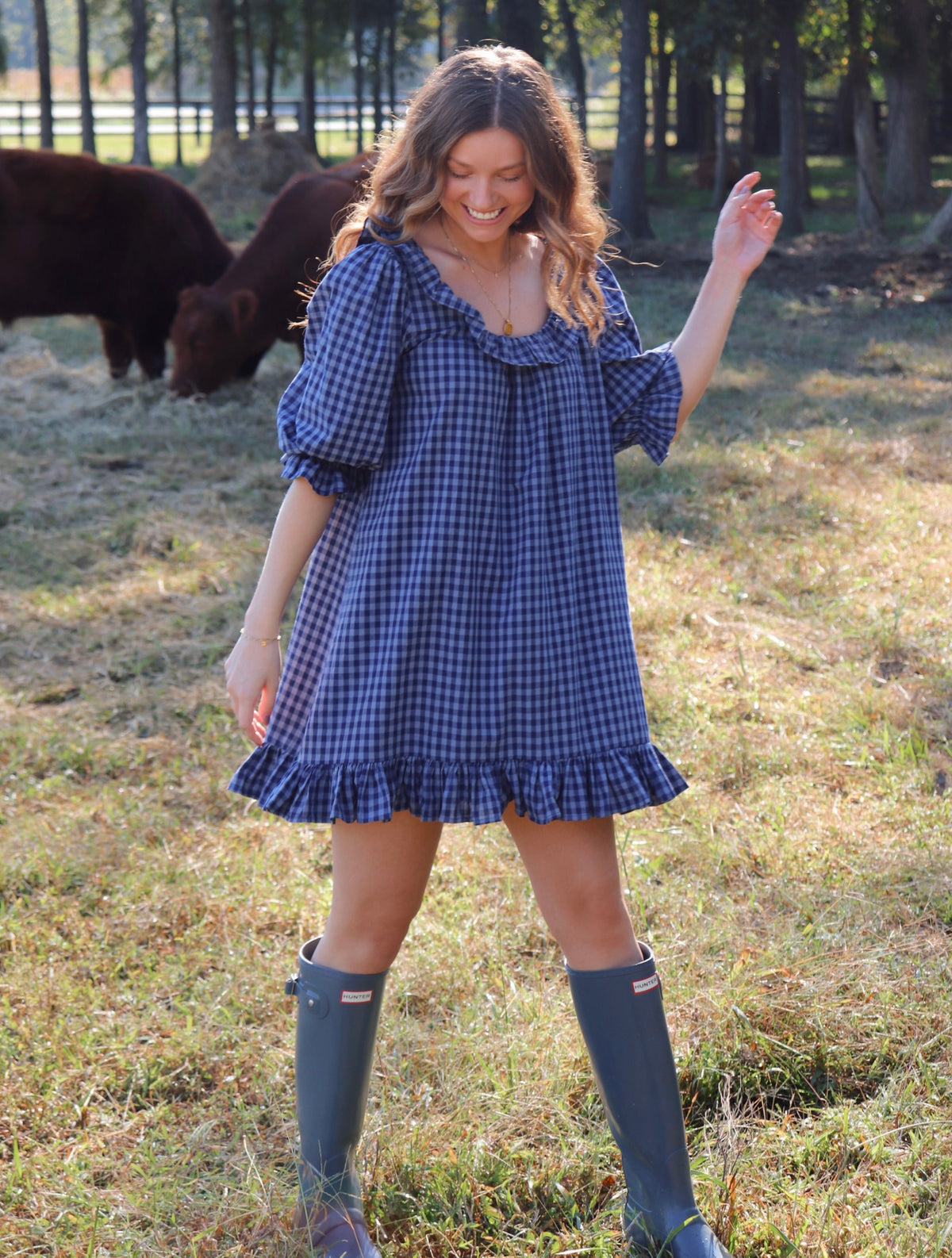 A woman wearing a blue gingham puff-sleeve dress with a ruffled neckline and hem, standing in a grassy field with cows in the background. She is wearing rain boots.