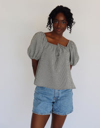 Woman wearing a black and white gingham blouse with puffed sleeves and denim shorts against a white background
