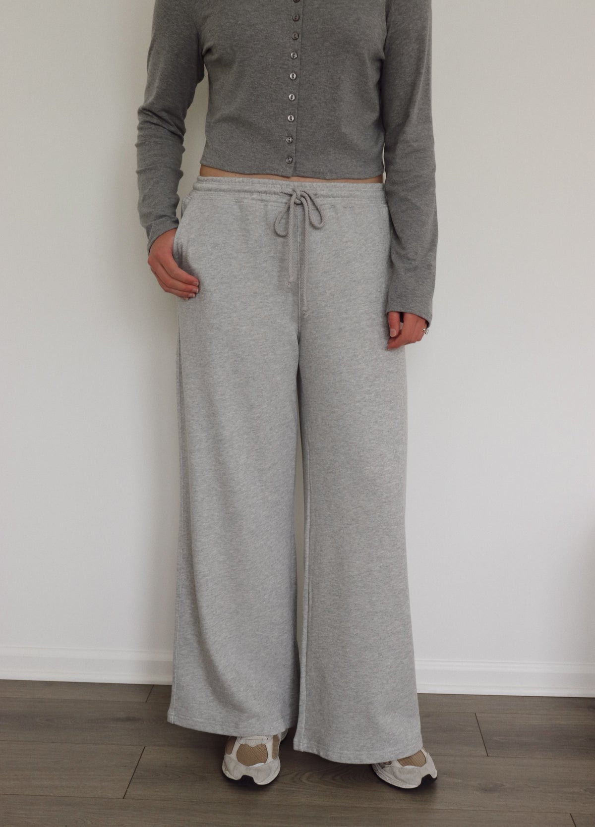 A person wearing light heather gray wide-leg sweatpants with a drawstring waist, paired with a gray long-sleeve cropped button-up top and beige sneakers, standing against a white wall.