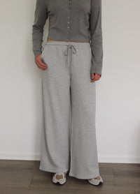 A person wearing light heather gray wide-leg sweatpants with a drawstring waist, paired with a gray long-sleeve cropped button-up top and beige sneakers, standing against a white wall.