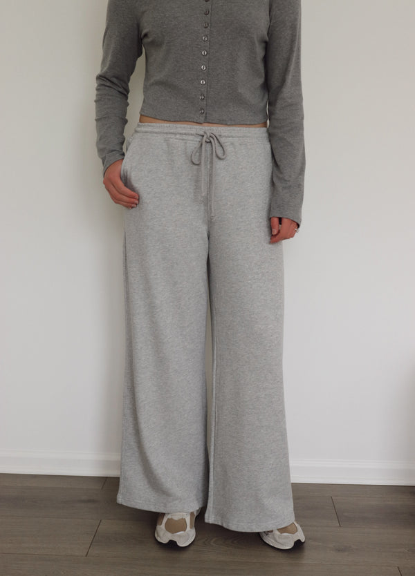 A person wearing light heather gray wide-leg sweatpants with a drawstring waist, paired with a gray long-sleeve cropped button-up top and beige sneakers, standing against a white wall.