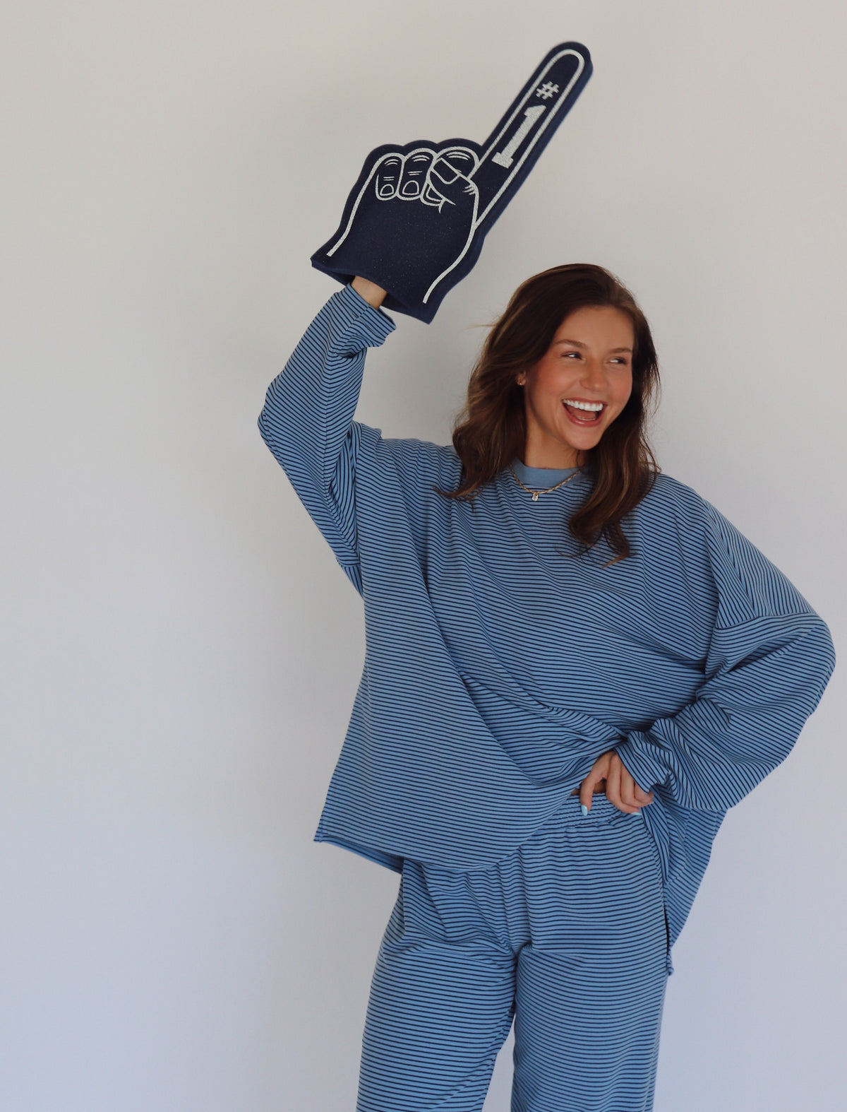 A woman modeling a matching blue striped lounge set, consisting of a long-sleeve top and wide-leg pants, against a plain white studio background while holding a navy blue foam finger that says #1