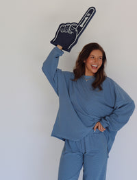 A woman modeling a matching blue striped lounge set, consisting of a long-sleeve top and wide-leg pants, against a plain white studio background while holding a navy blue foam finger that says #1