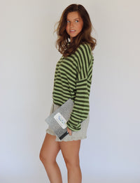 A woman modeling a green and dark green striped long-sleeve button-up cardigan with light beige ruffled shorts, holding a black and white patterned notebook against a plain white background.