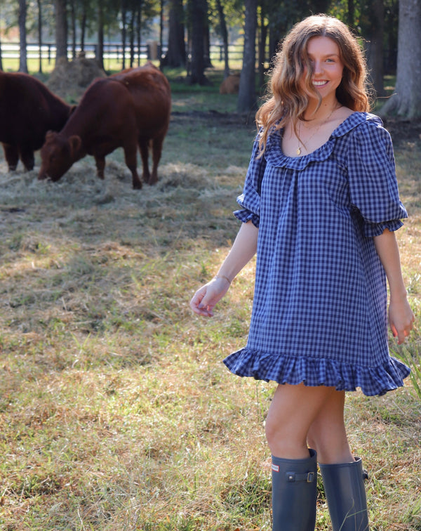 A woman wearing a blue gingham puff-sleeve dress with a ruffled neckline and hem, standing in a grassy field with cows in the background. She is wearing rain boots.