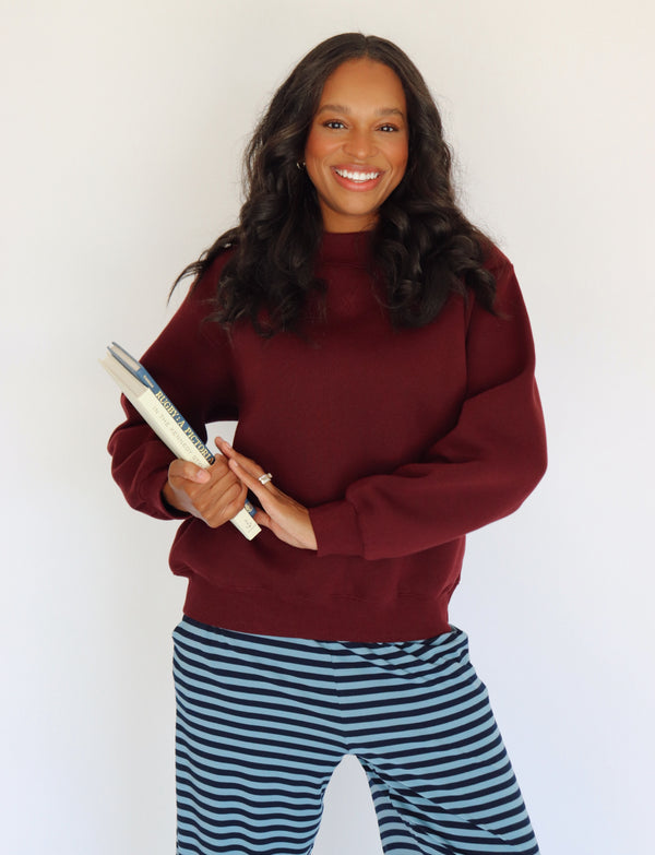 A woman wearing a burgundy mock neck pullover and blue-and-white striped pants holds books against a plain white background.