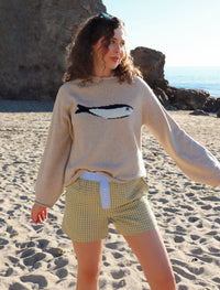 Woman wearing a beige sweater with a fish design and checkered shorts on a beach.