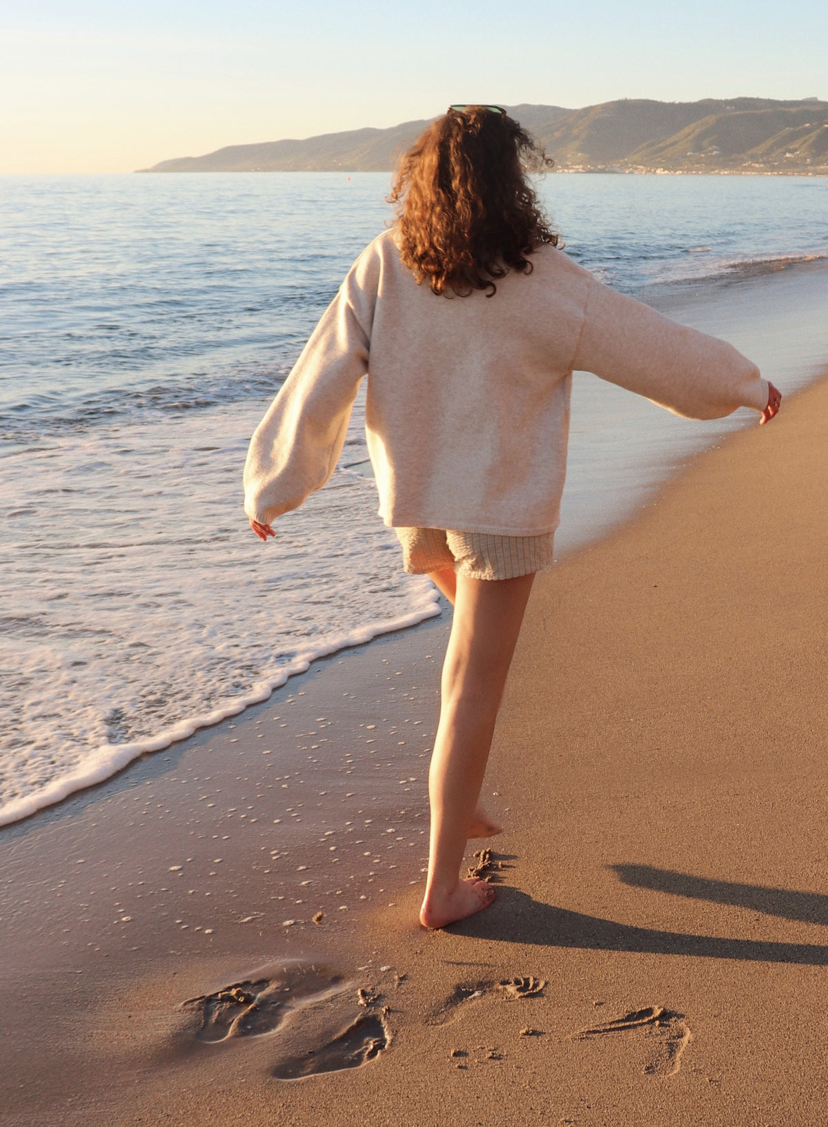 Woman in a beige cardigan and shorts walking on a sandy beach with ocean in the background