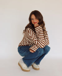 Woman wearing a brown and white striped sweater cardigan and blue jeans sitting on a white background