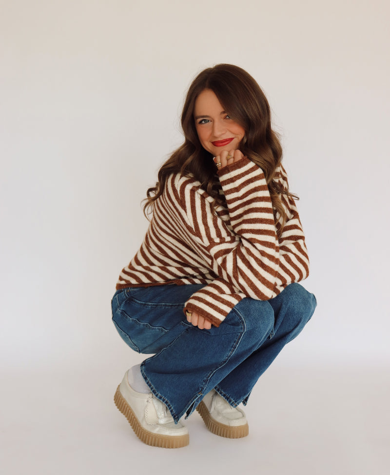 Woman wearing a brown and white striped sweater cardigan and blue jeans sitting on a white background