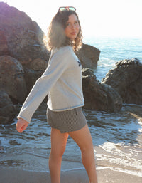 Woman standing on a rocky beach wearing a light sweater and plaid shorts.