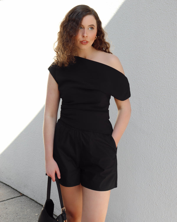 Woman wearing a black off-shoulder top and shorts against a light gray wall.