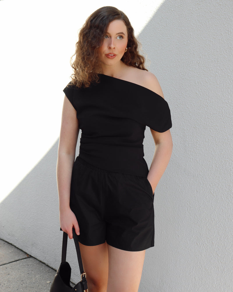 Woman wearing a black off-shoulder top and shorts against a light gray wall.