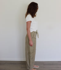 A woman stands in profile wearing a white short-sleeve t-shirt and high-waisted, wide-leg khaki cargo pants with a side tie, set against a plain white wall and light wood floor.