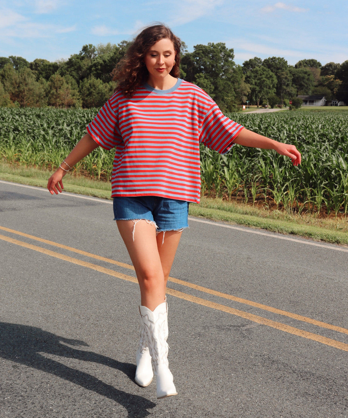 Woman in a striped shirt and denim shorts standing on a road with a cornfield in the background