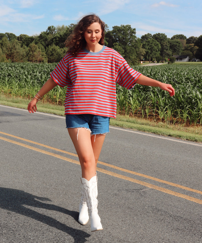 Woman in a striped shirt and denim shorts standing on a road with a cornfield in the background