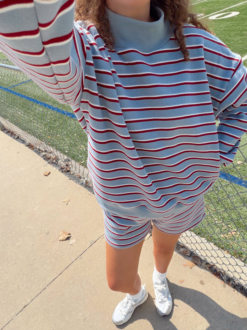 A person outdoors on a sports field wearing an oversized light blue striped sweatshirt and matching shorts with red and white horizontal stripes, paired with white sneakers.