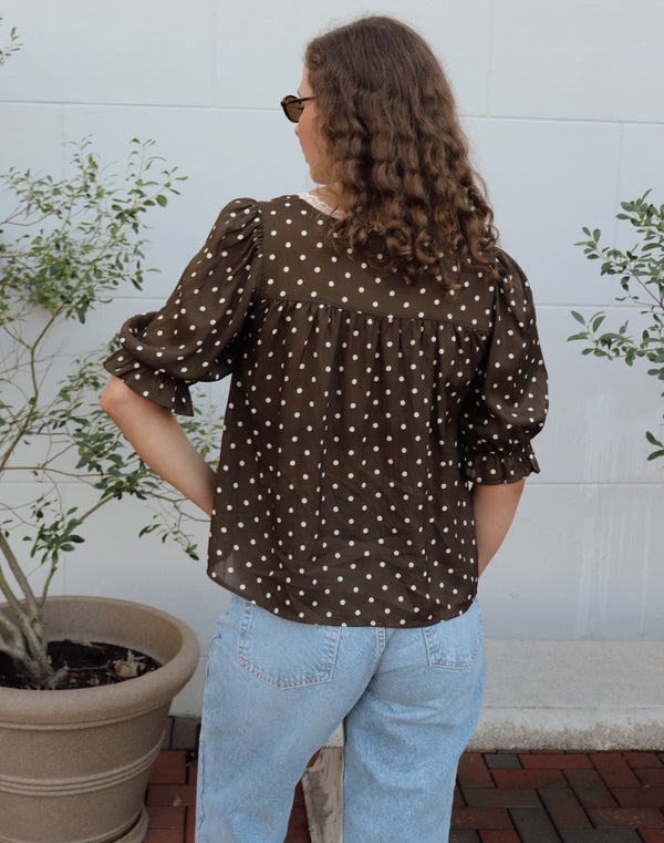 Woman wearing a brown polka dot blouse and blue jeans standing outdoors with plants in the background.