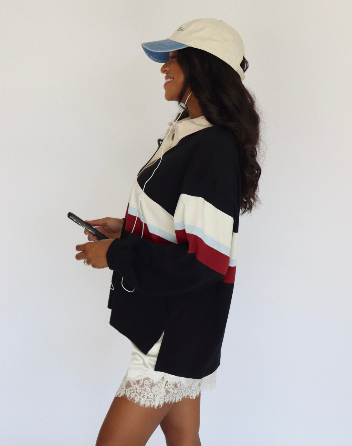 A woman in profile wearing a black, white, and red color-block hoodie, a cream baseball cap, and white lace shorts, holding a phone against a plain white background.