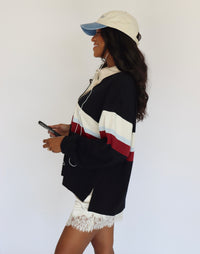 A woman in profile wearing a black, white, and red color-block hoodie, a cream baseball cap, and white lace shorts, holding a phone against a plain white background.