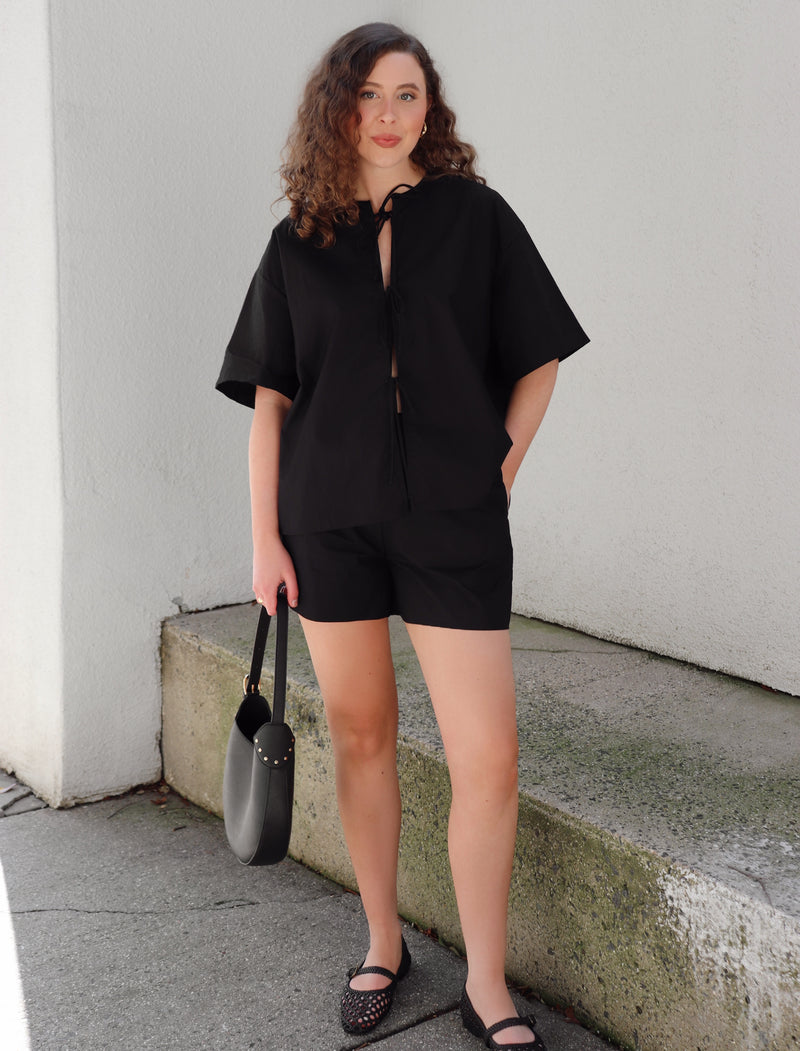 Woman in a black outfit standing on concrete background against a beige  wall.