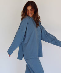 A woman modeling a matching blue striped lounge set, consisting of a long-sleeve top and wide-leg pants, against a plain white studio background.