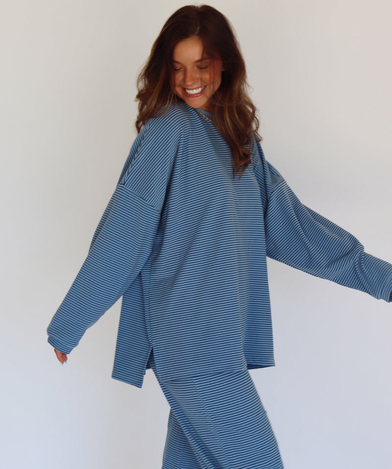 A woman modeling a matching blue striped lounge set, consisting of a long-sleeve top and wide-leg pants, against a plain white studio background.