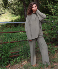 A woman wearing a matching olive green striped lounge set stands outdoors in a wooded area with greenery and a rustic fence behind her.