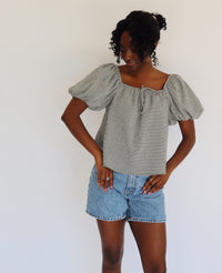 Woman wearing a black and white gingham blouse with puffed sleeves and denim shorts against a white background