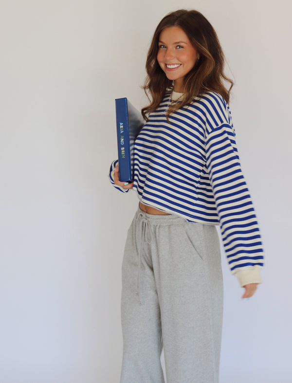 A woman smiling and holding a blue book, wearing a blue and white striped long-sleeve crop top and light gray sweatpants against a plain white background.