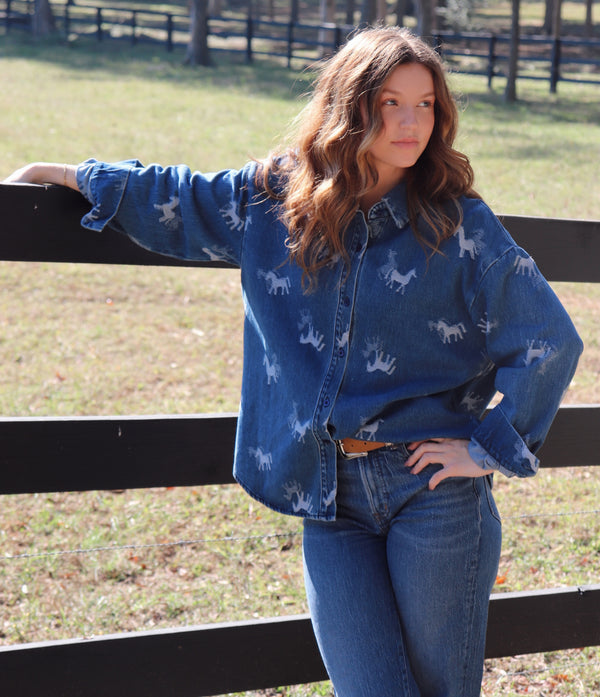 A woman wearing a blue denim button-up shirt with a white horse print, leaning against a black wooden fence in an outdoor field.