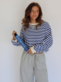 A woman modeling a blue and white striped long-sleeve crop top with cream ribbed cuffs, paired with light gray sweatpants, holding a blue binder against a plain white background.