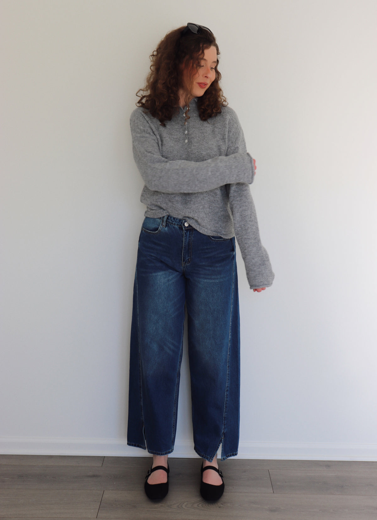 Woman wearing a gray sweater top and blue jeans standing against a white wall.