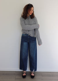 Woman wearing a gray sweater top and blue jeans standing against a white wall.