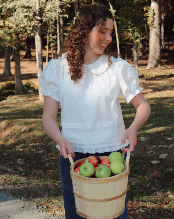 A woman with curly brown hair stands outdoors in a wooded area holding a wooden basket filled with green and red apples. She is wearing a white short-sleeve blouse with a ruffled collar and dark blue pants.