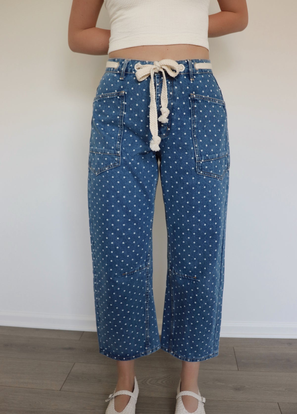 woman wearing white tank top and blue jeans with white polka dots on them against a white wall