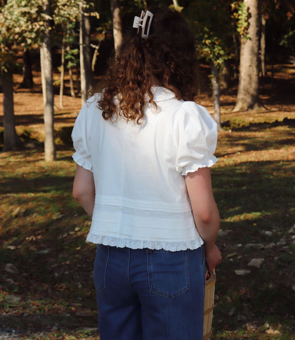 A person viewed from behind wearing a white short-sleeve puff-sleeve blouse with a ruffled hem and blue jeans, standing outdoors in a wooded area.