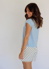 A woman modeling a light blue short-sleeve button-up shirt and white shorts with black polka dots, standing against a plain light background.