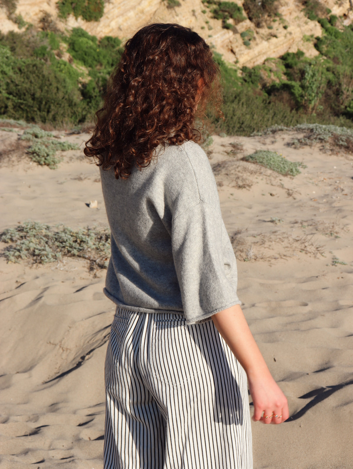 Person wearing a gray top and striped pants standing on a sandy beach.