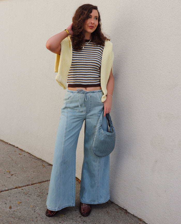 Woman wearing a striped shirt, a yellow sweater around her shoulders  and blue jeans against a gray wall.