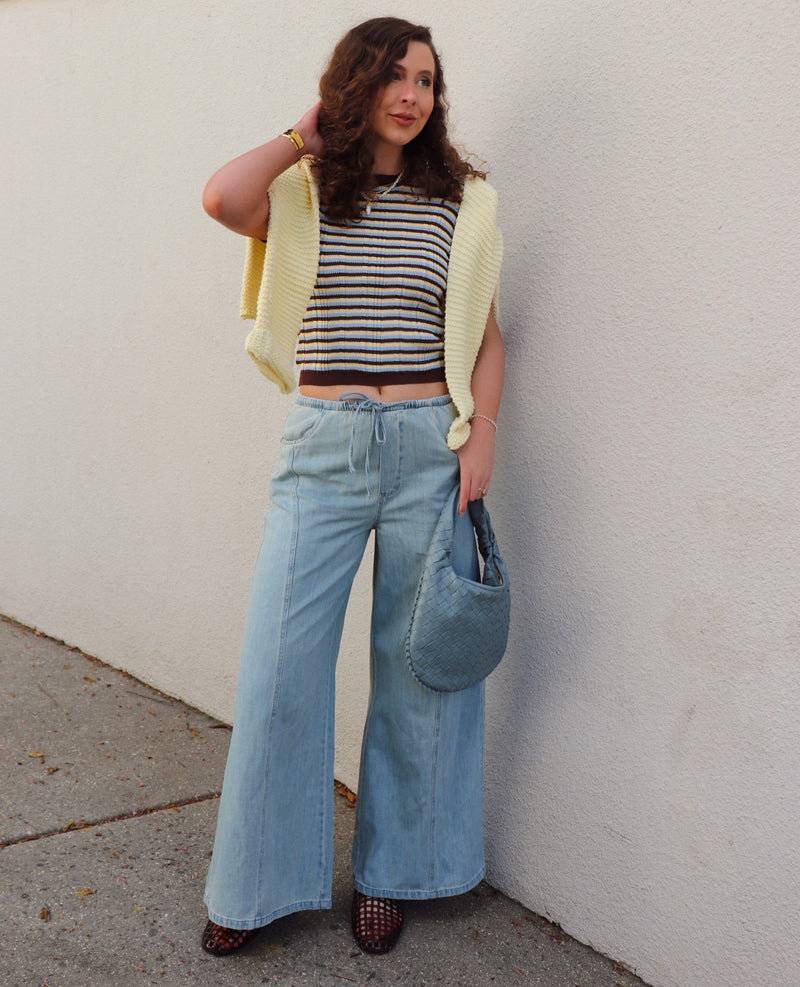 Woman wearing a striped shirt, a yellow sweater around her shoulders  and blue jeans against a gray wall.